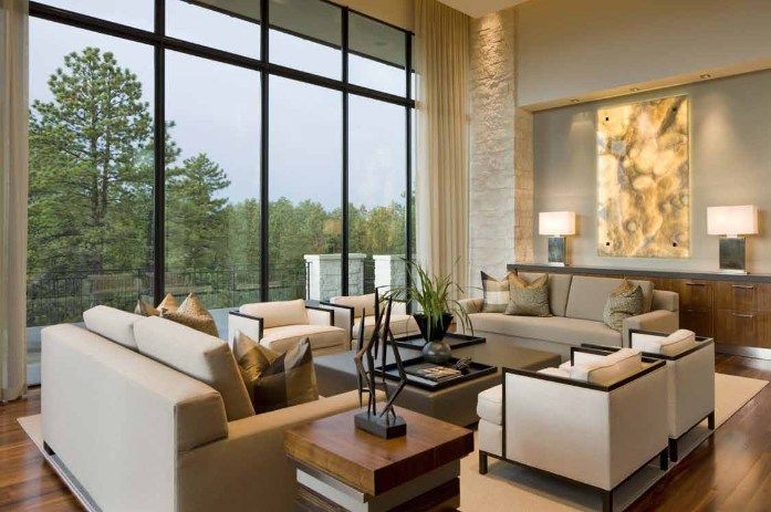 Living room with large windows overlooking trees; beige sofas, wood furniture, neutral color scheme.