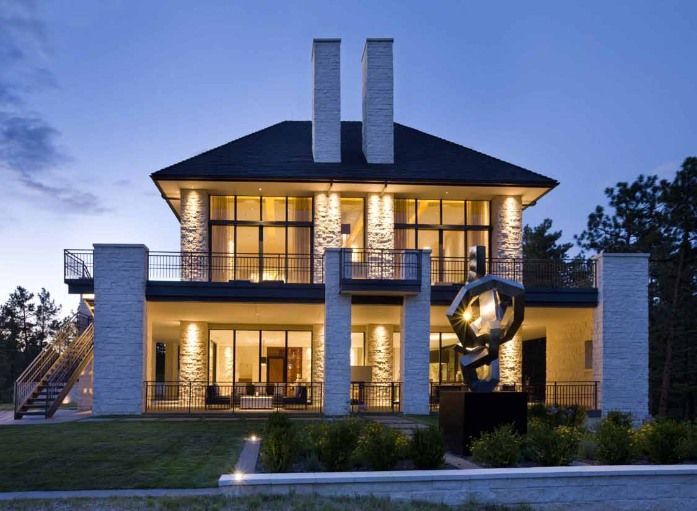 Modern two-story house with stone facade, large windows, and outdoor sculpture at dusk; lit by warm interior lights.
