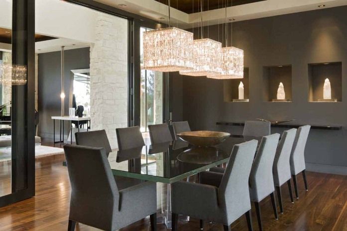 Elegant dining room with glass table, gray chairs, dark walls, and rectangular crystal chandeliers.