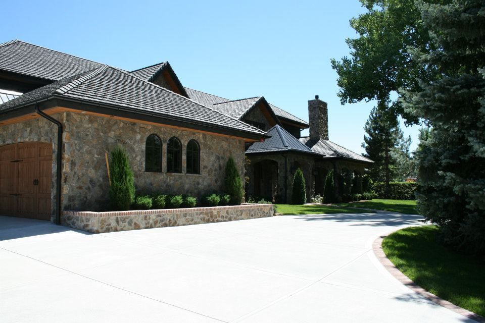 Stone-faced house with a long driveway and garage, set against a blue sky, with trees in the foreground.