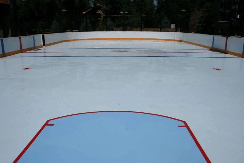 Empty ice hockey rink, blue and red markings, white boards, trees in background.