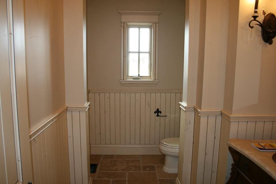 Small, cream-colored bathroom with toilet, wainscoting, a small window, and stone tile flooring.