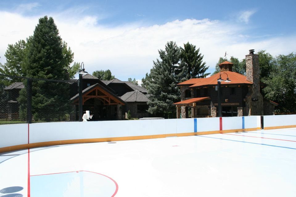 Outdoor ice rink in front of two-story houses with brown roofs and stone accents, blue sky.
