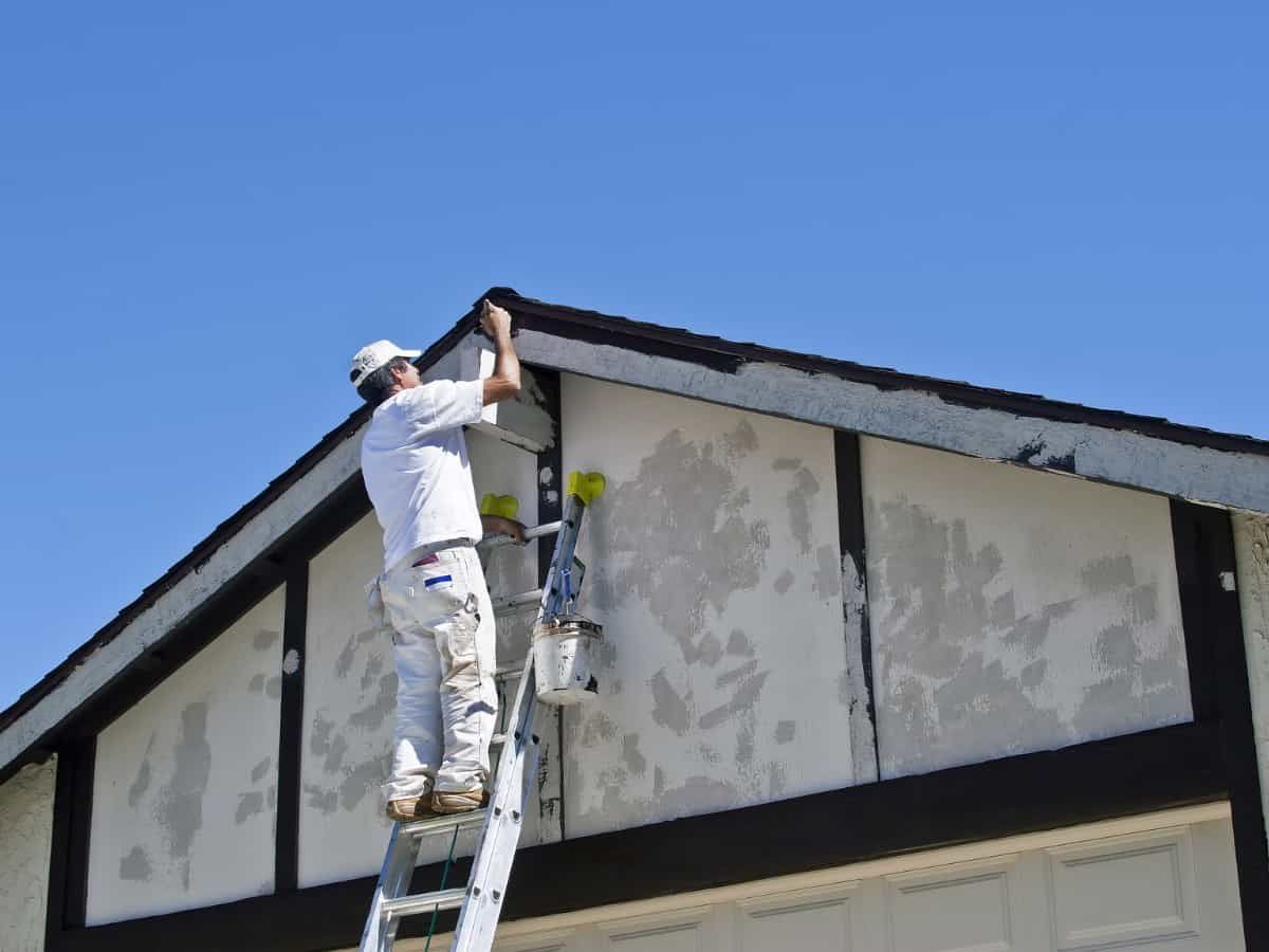 A worker in white clothing stands on a ladder, painting the trim on a house exterior against a clear blue sky.