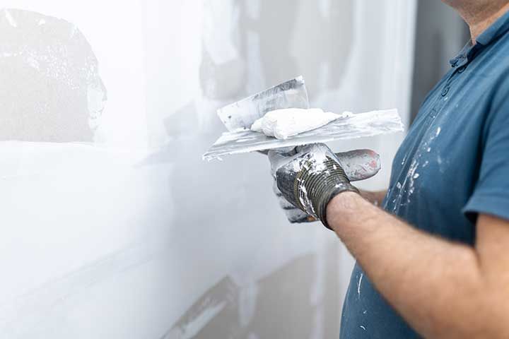 A person wearing a blue shirt and work gloves uses a metal putty knife to apply joint compound to a white wall.