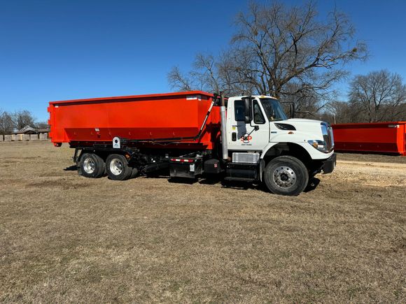 A dump truck is parked in a field with trees in the background.