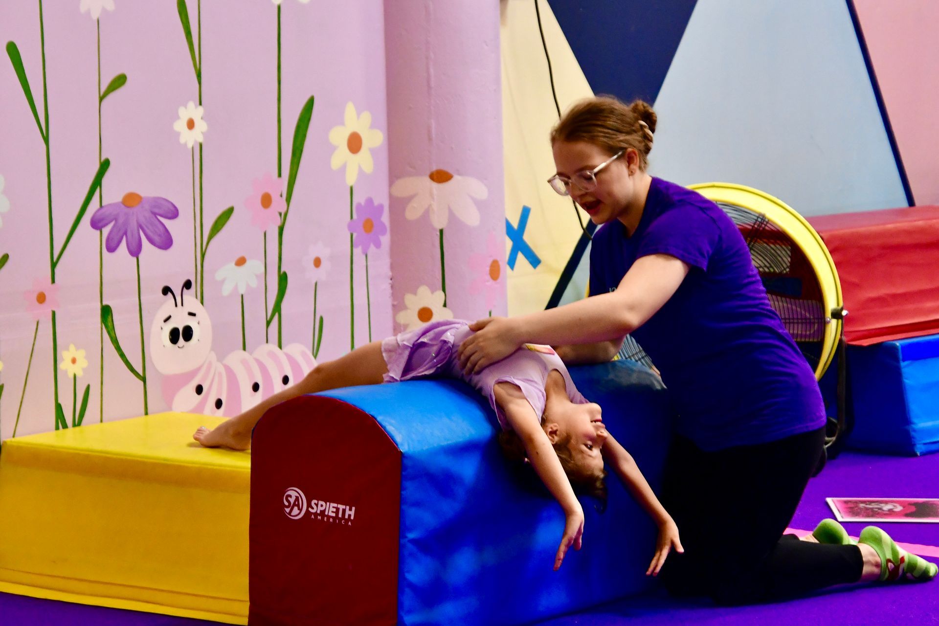 A preschool coach assisting a preschooler attempt a backbend over the boulder