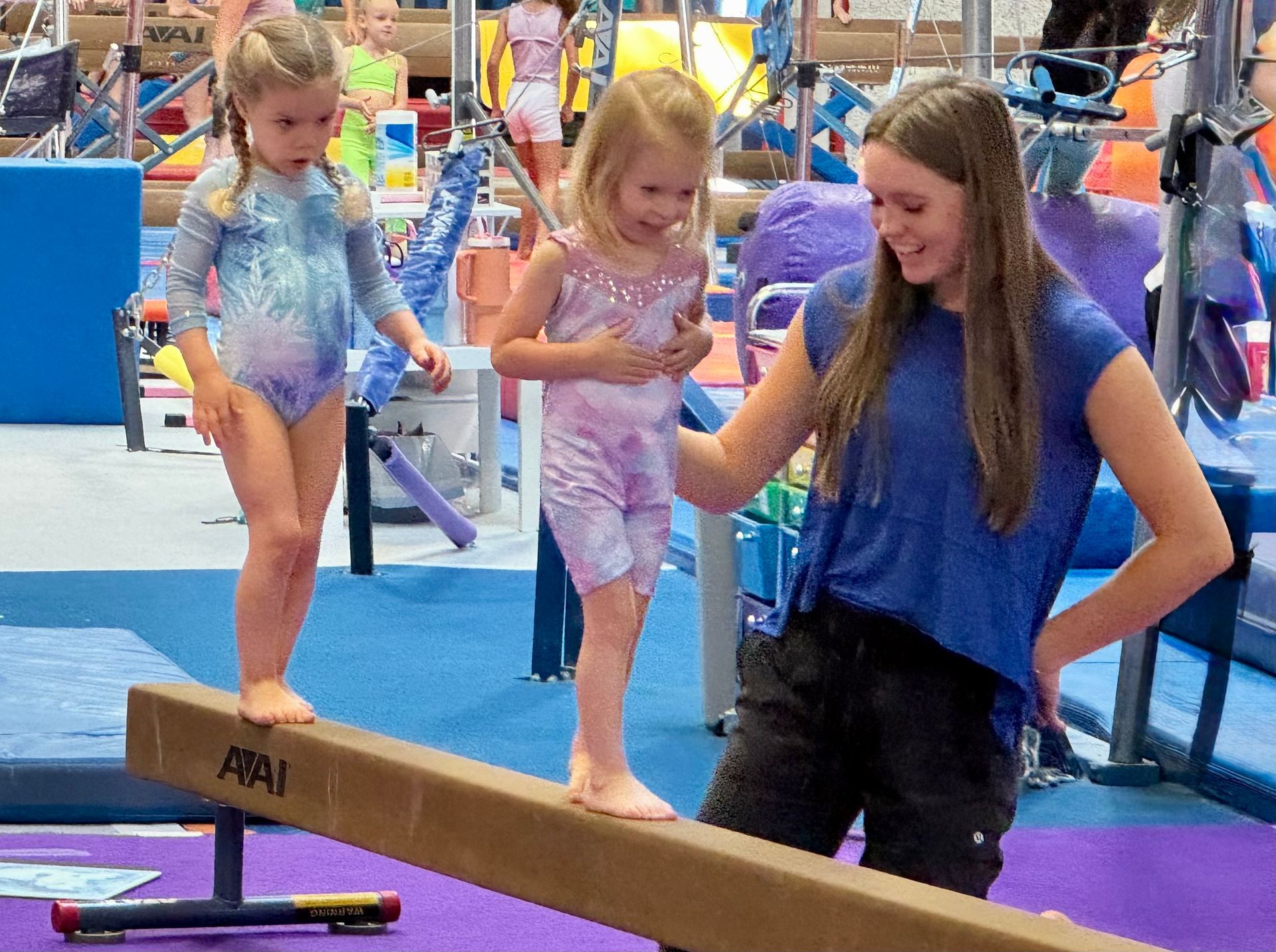 Smiling preschool coach helping two young girls in leotards on balance beam