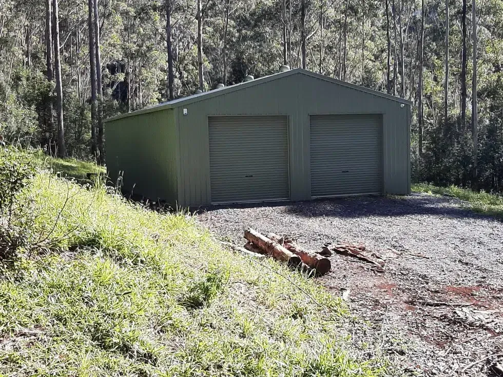 A green garage is sitting on top of a grassy hill in the middle of a forest.