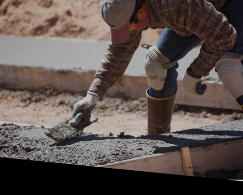 Worker smoothing wet concrete at a construction site, wearing gloves and boots.