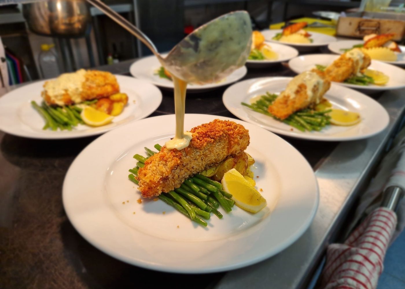 Crusted fish served with greens and lemon while sauce is poured, reflecting fresh, nutritious and e