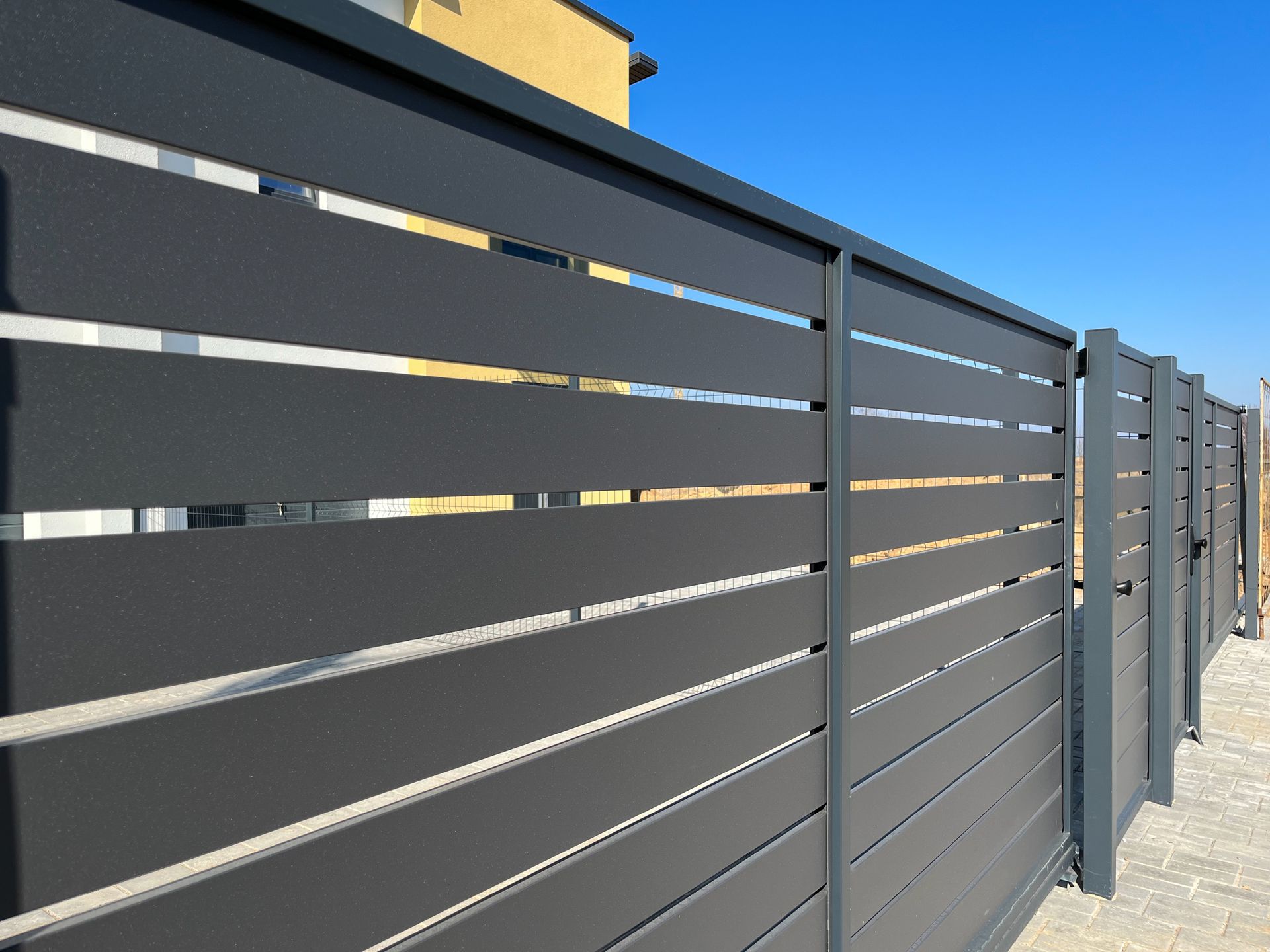 Dark gray slatted metal fence along a paved walkway under a bright sky Dark gray slatted metal fence along a paved walkway under a bright sky