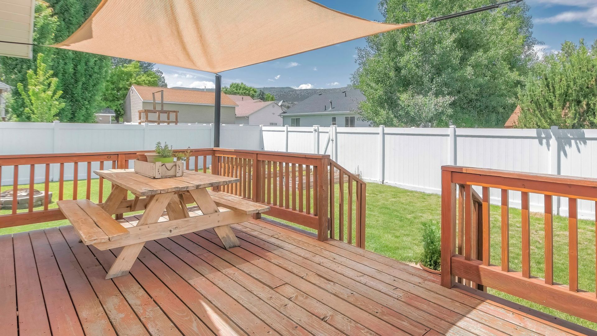 Wooden deck with picnic table, beige umbrella, and green yard surrounded by white fencing