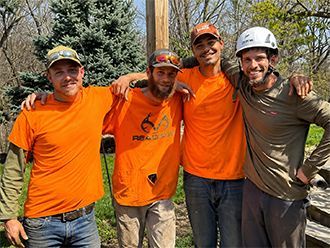 Four workers in orange shirts and hard hats stand arm-in-arm at a construction site, smiling.