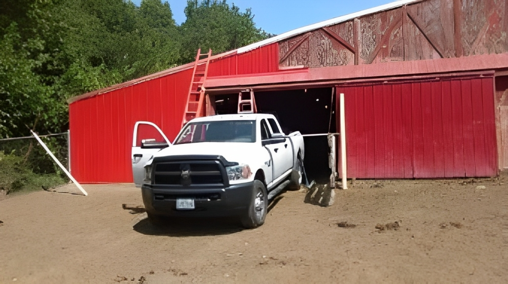 White pickup truck parked in front of a red barn with open doors on a dirt yard