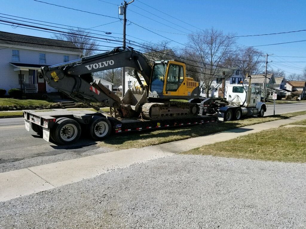 Yellow and black Volvo excavator on a flatbed trailer, being towed by a white semi-truck on a residential street.