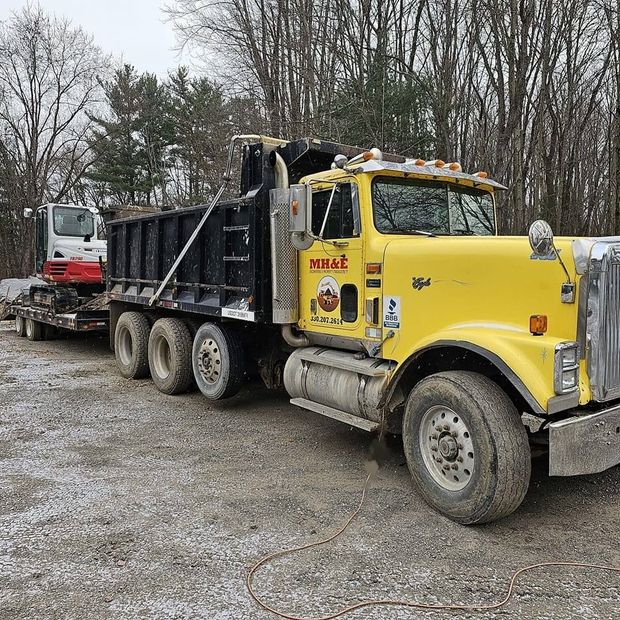 Yellow dump truck with a black bed and white excavator on a trailer in a gravel lot.