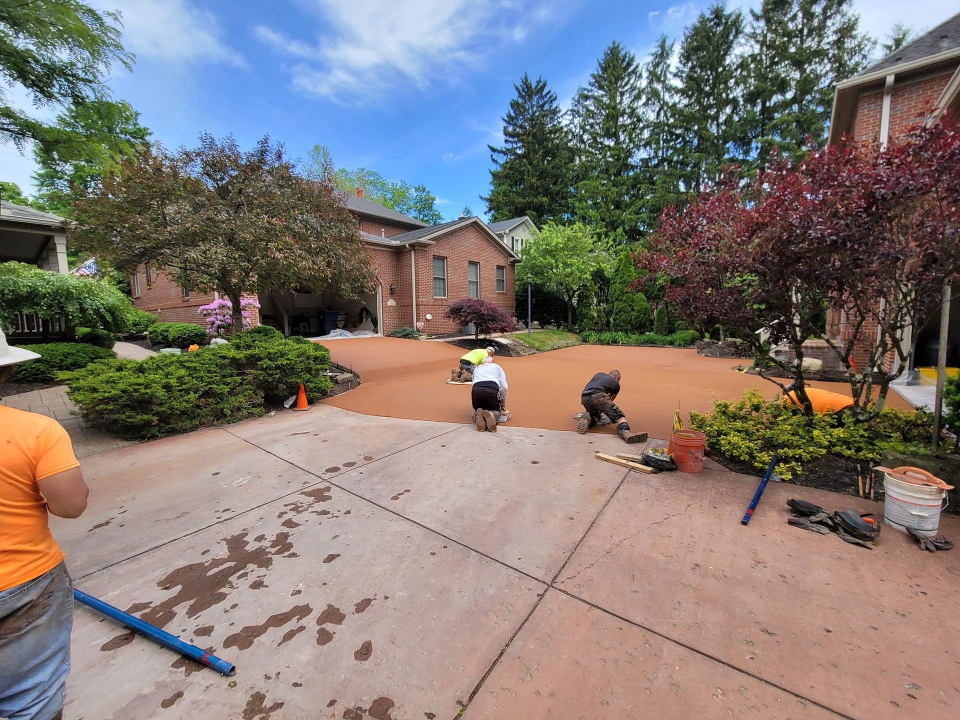 Workers laying mulch in a backyard with a brick house and surrounding trees under a blue sky.