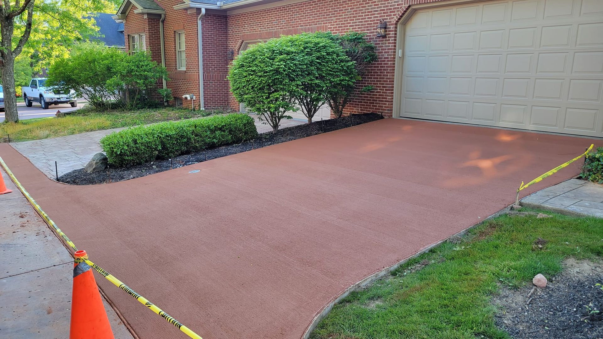Newly paved red-brown driveway with a green lawn, bushes, and a brick house with a garage.