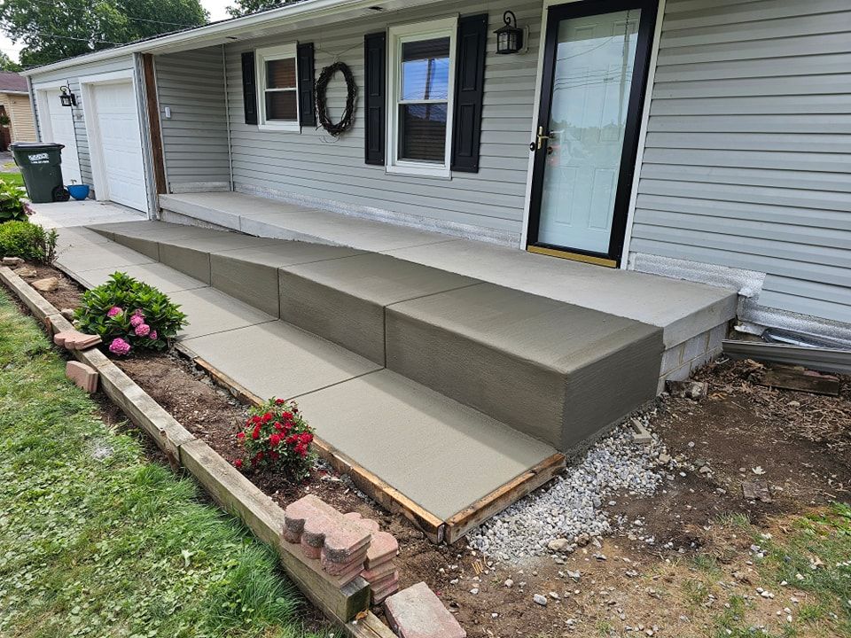 New concrete ramp and steps leading to a house entrance. Green grass and garden beds in foreground. Gray siding.