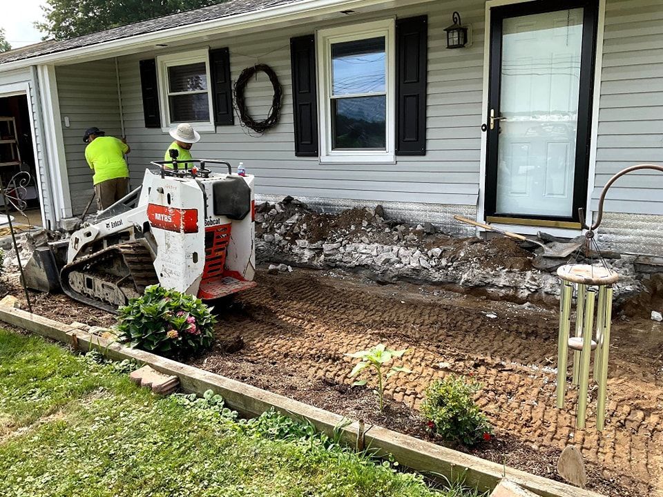 Construction worker operating Bobcat excavator, removing dirt near a house with black shutters and a white door.