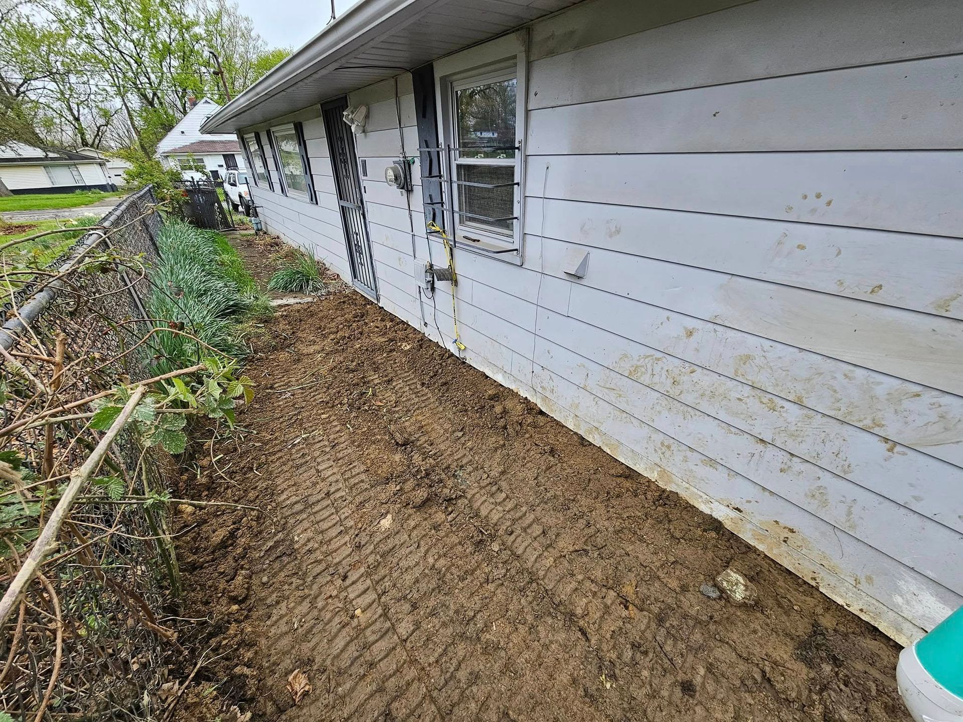 Muddy ground next to a white house with a small window. A fence with foliage is to the left.