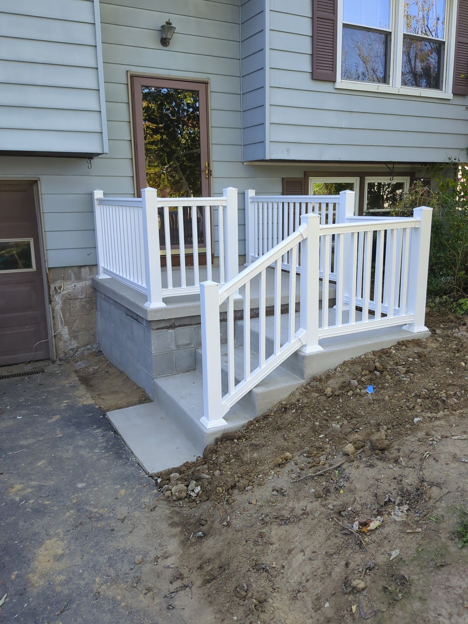 White railing surrounds a concrete porch with steps leading to a house entrance. The house has gray siding and brown shutters.