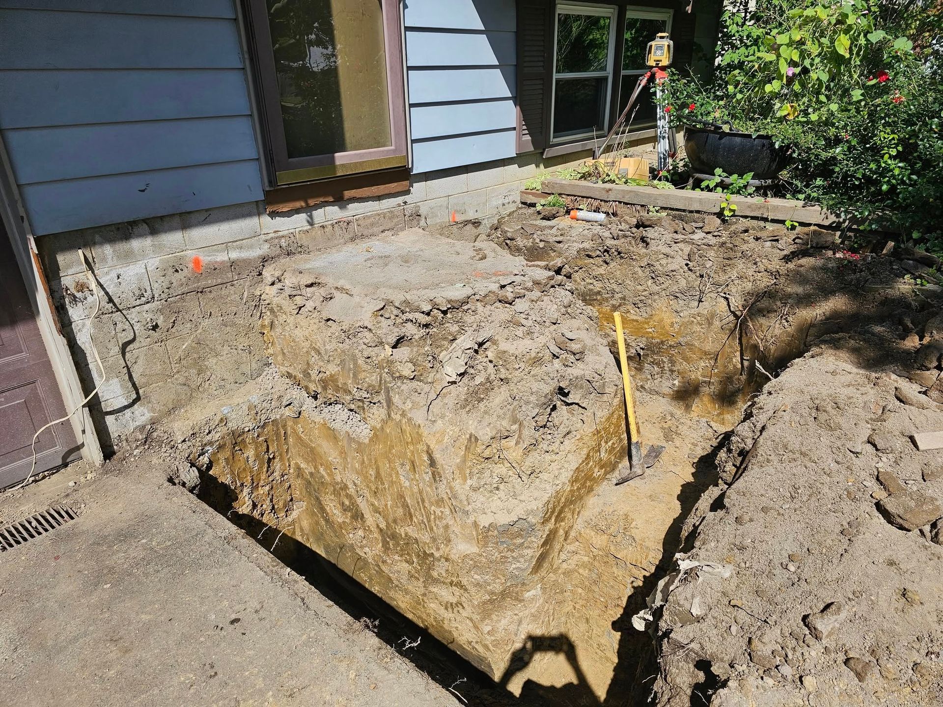 Excavation next to a house with a concrete foundation; dirt piles and a shovel visible.