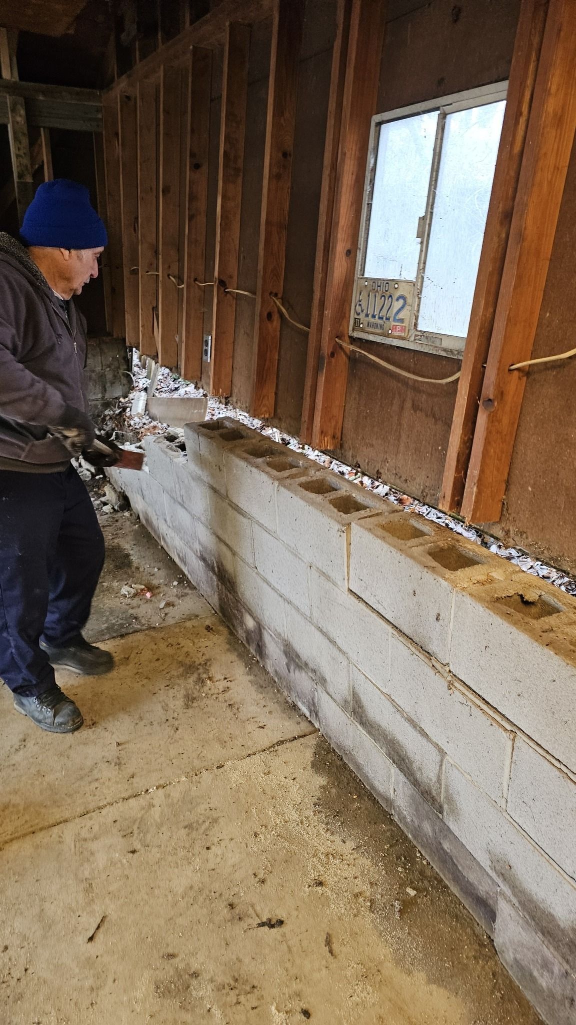 Person working on concrete block wall next to window and wall studs; interior setting.