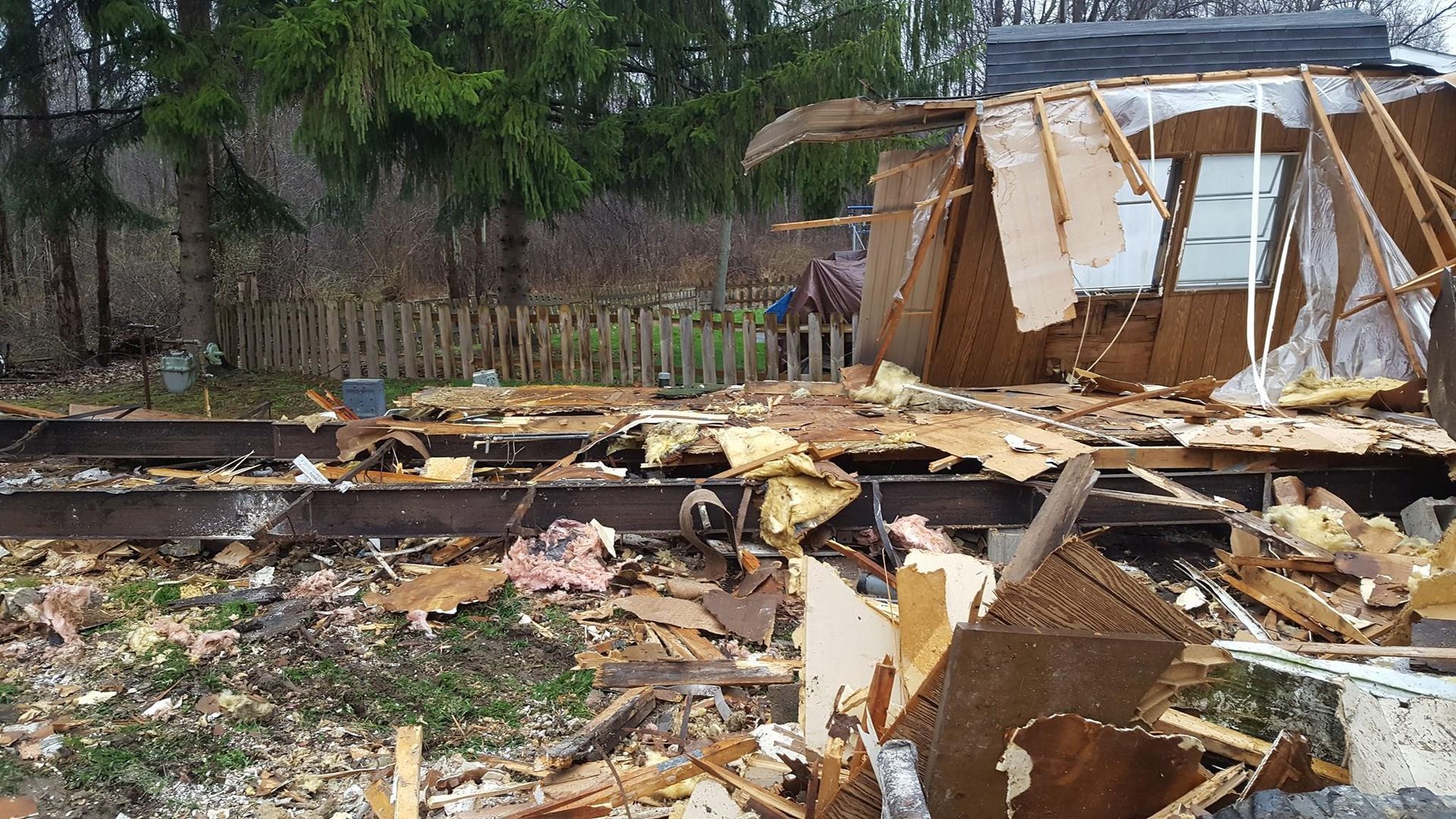 Debris of demolished building; wooden structure and rubble on ground. Fence and trees in background.