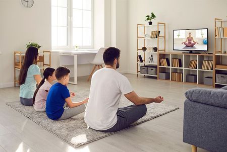 A family is sitting on the floor in a living room watching a yoga video on tv.