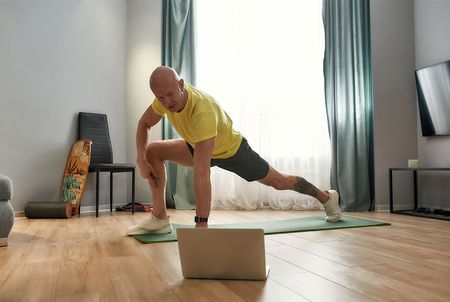 A man is stretching on a yoga mat in front of a laptop computer.