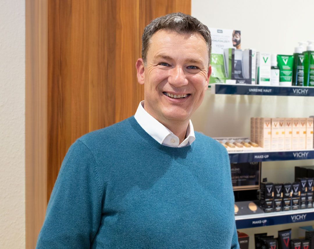 Ein Mann in einem blauen Pullover lächelt vor einem Regal mit Kosmetika.