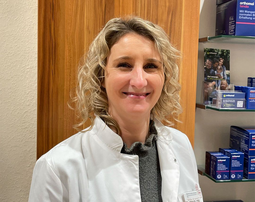 Eine Frau in einem weißen Laborkittel lächelt vor einer Holzwand.