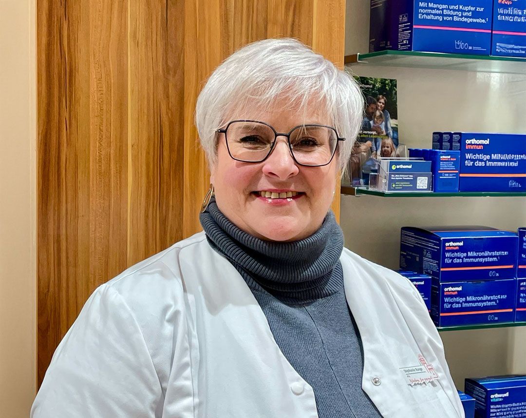 Eine Frau mit Brille und weißem Kittel steht vor einem Regal.