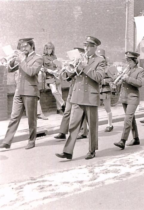 Een fanfare marcheert door een straat en speelt op instrumenten. De muzikanten in uniform dragen trompetten en het lijkt alsof het zich in de open lucht afspeelt.