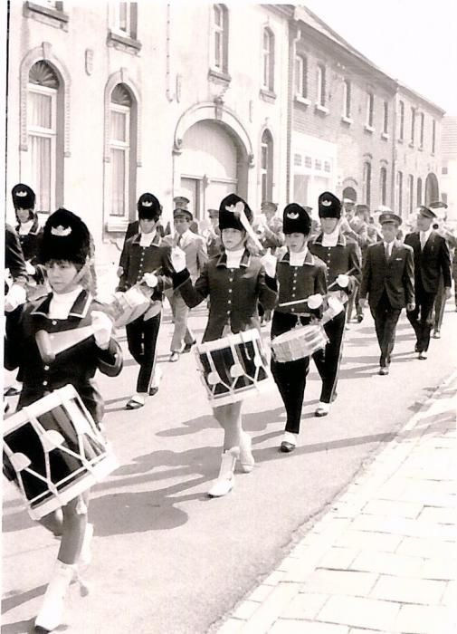 Een fanfare, in uniform, speelt op trommels en marcheert door een straat met gebouwen.
