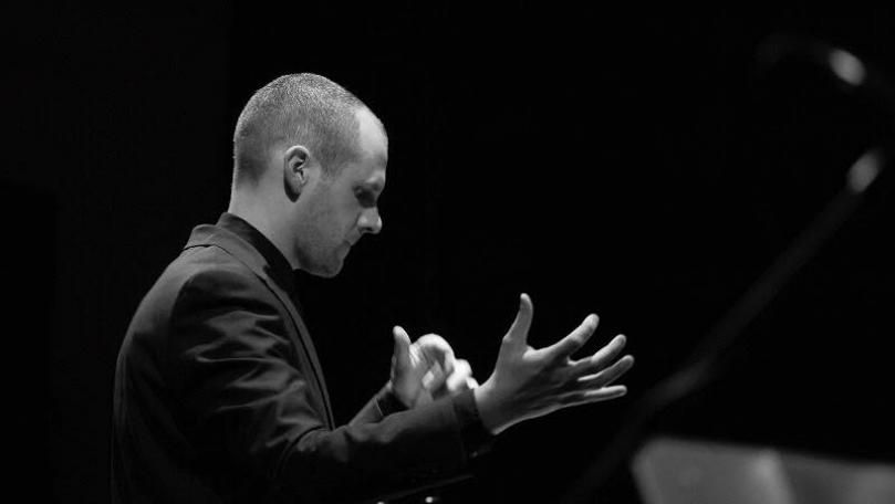 Een man zit achter een piano met zijn handen omhoog en kijkt naar beneden in een donkere omgeving, waarschijnlijk tijdens een optreden.