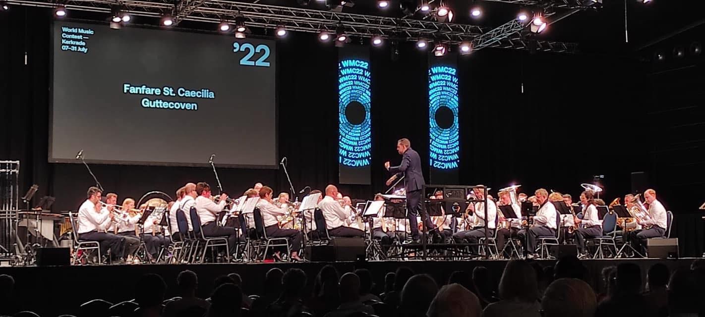 Een orkest treedt op een podium op, een dirigent geeft leiding. Op een scherm verschijnt het jaartal '22 en tekst. Publiek op de voorgrond.