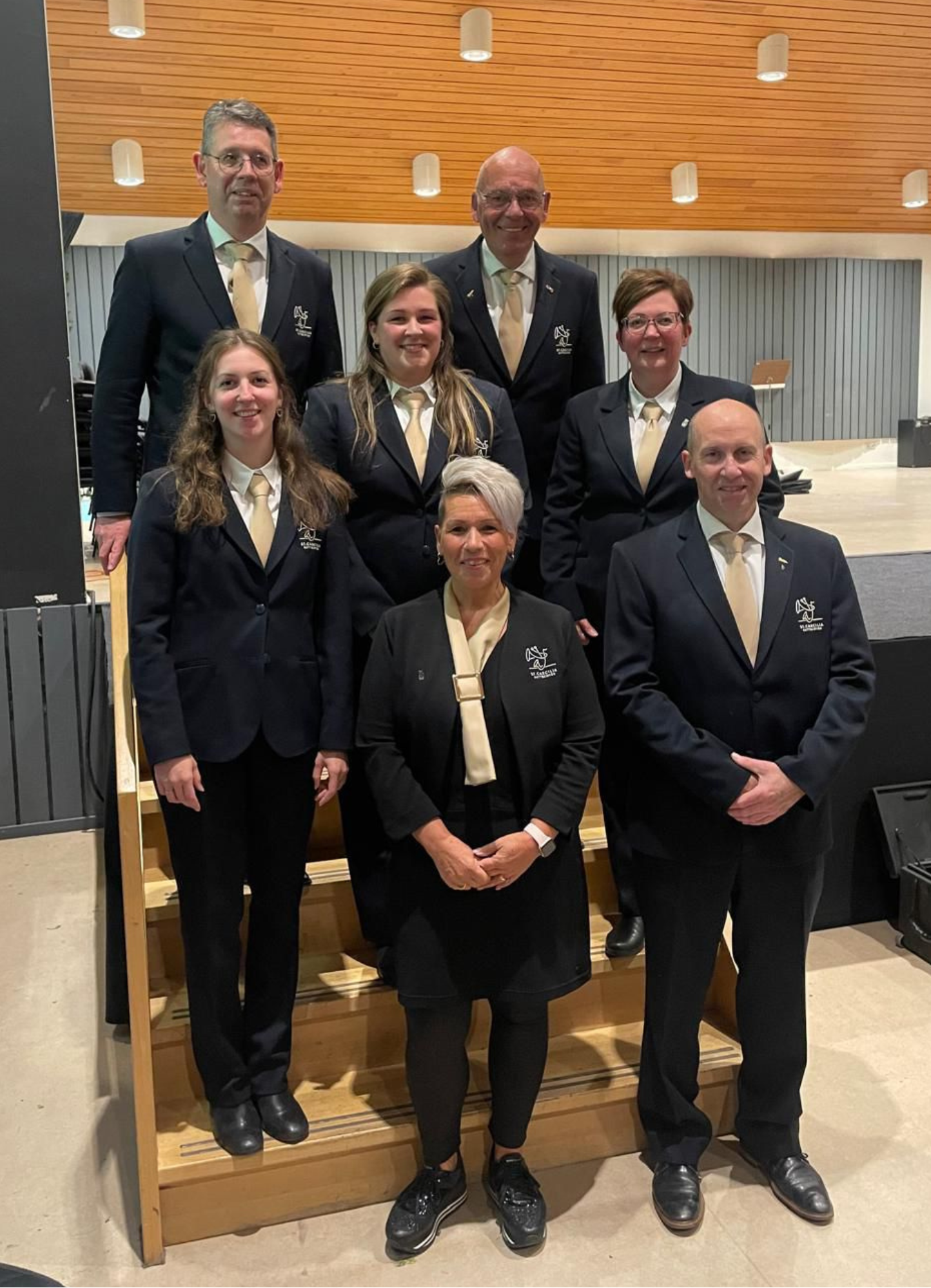 Een groep mensen in bijpassende donkere pakken en beige stropdassen poseert op de trappen van een hal.