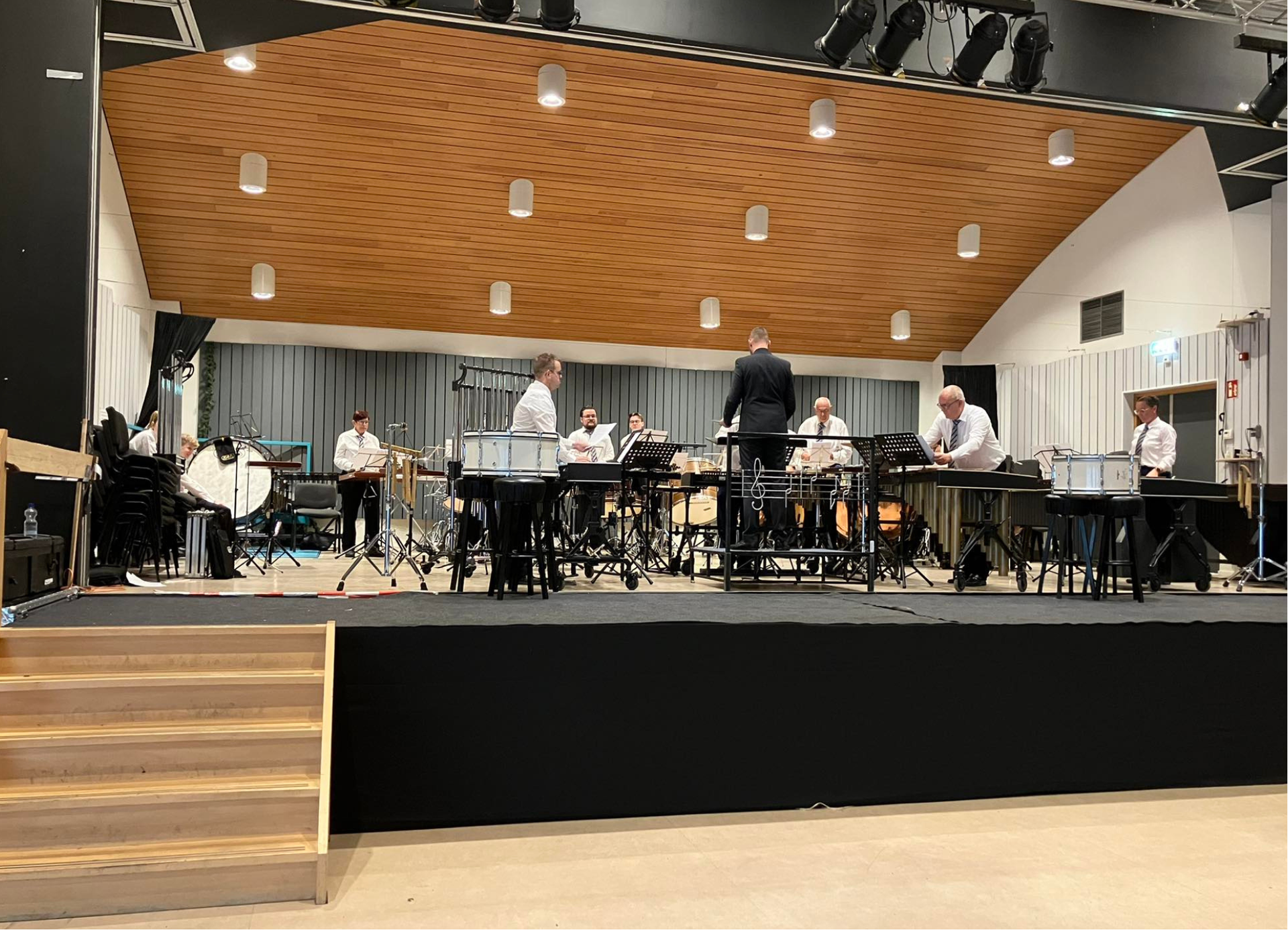 Een muzikaal ensemble treedt op een podium op. Musici in witte overhemden bespelen instrumenten onder de lampen in een zaal met houten lambrisering.