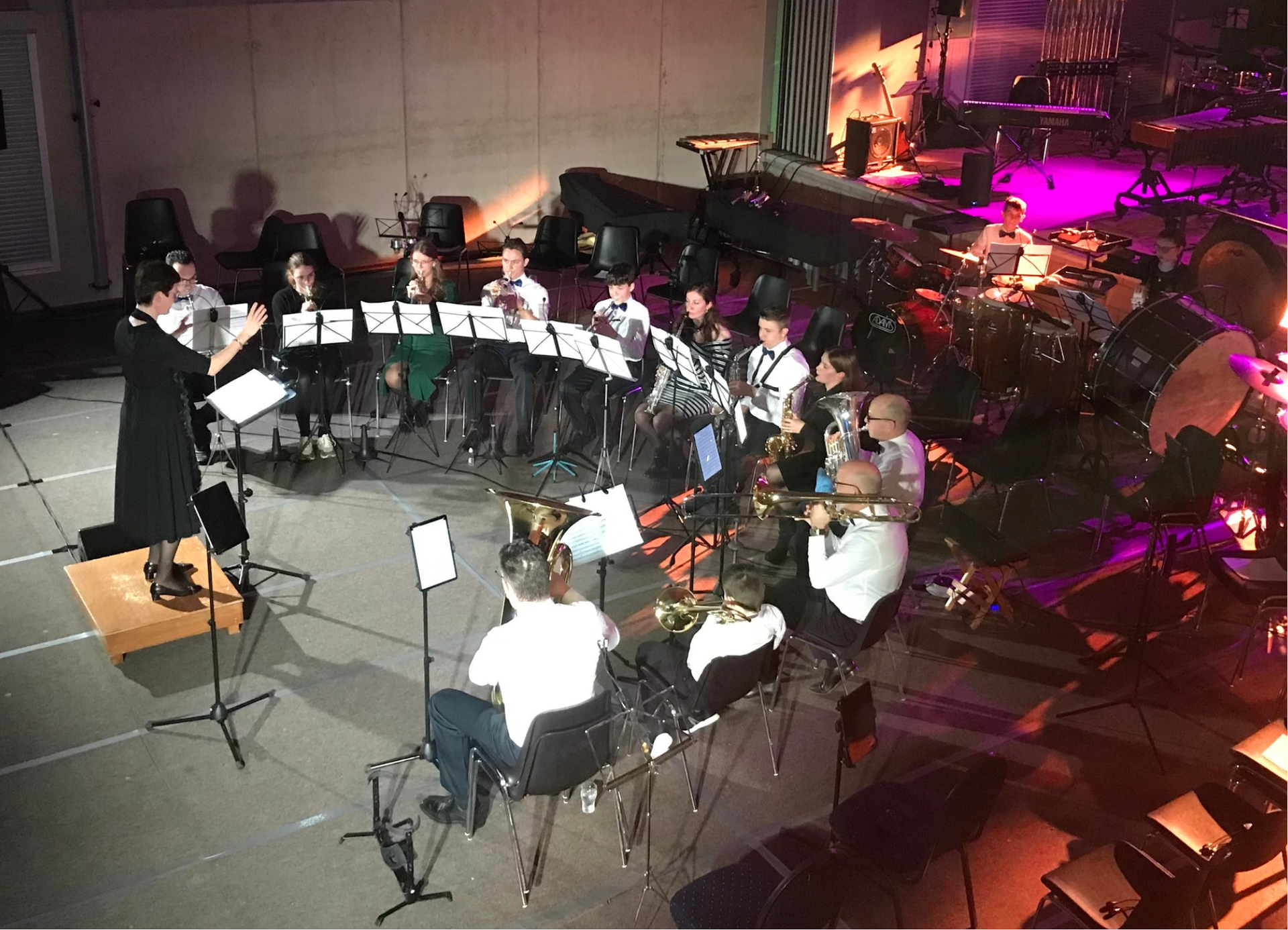 Een orkest repeteert op het podium. De dirigent staat tegenover het ensemble, de muzikanten lezen de bladmuziek, en er zijn drums en andere instrumenten aanwezig.