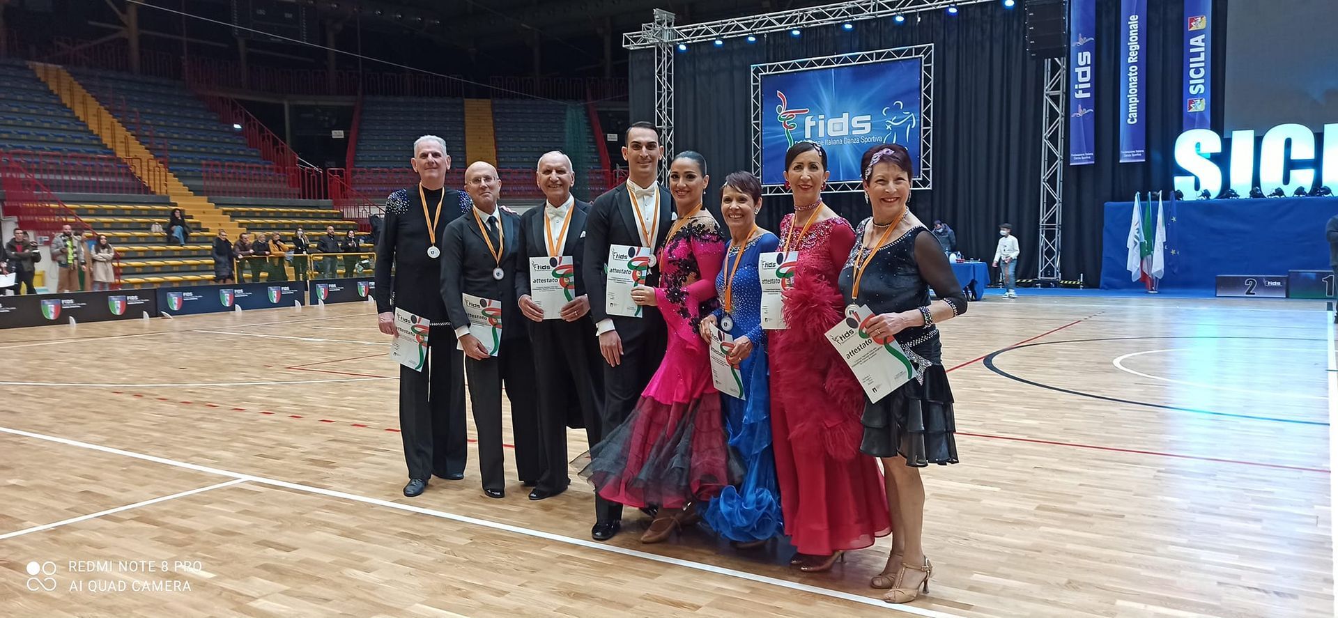 Gruppo di persone in abiti formali, probabilmente una gara di ballo. Ambientazione: arena con un palco sullo sfondo.