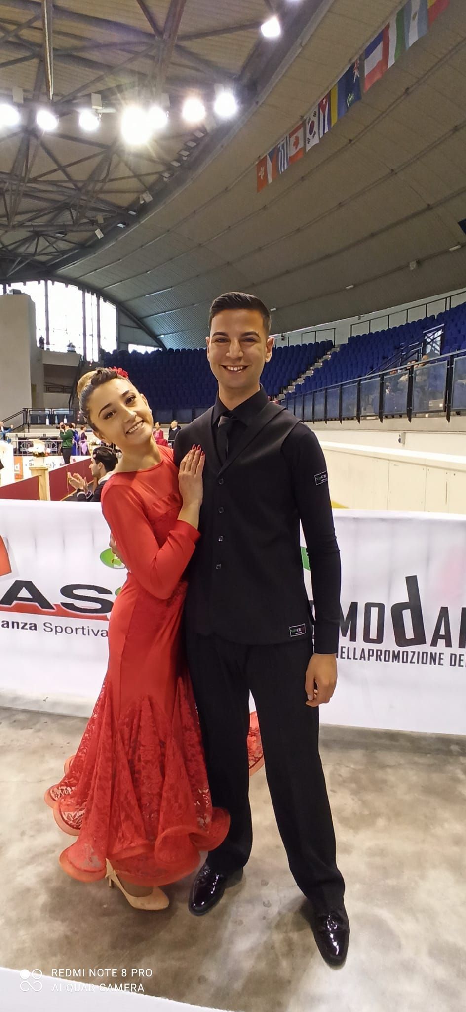 Una coppia di ballerini posa in un'arena. La donna indossa un abito rosso e sorride. L'uomo è vestito di nero, sorridente, con un braccio intorno alla vita.