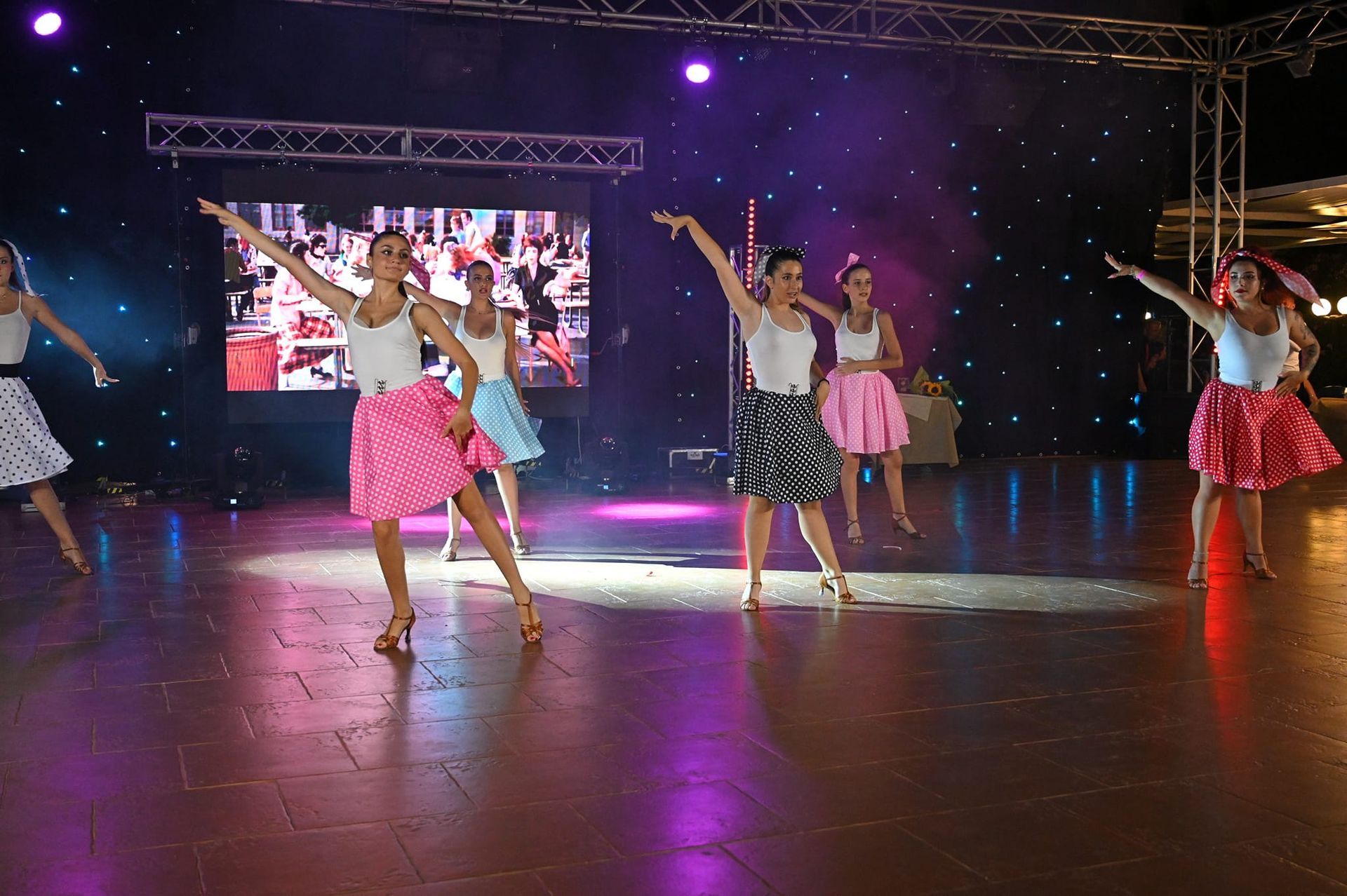 Ballerini sul palco in abiti in stile anni '50, con le braccia alzate. Gonne colorate, top bianchi, davanti a uno schermo.