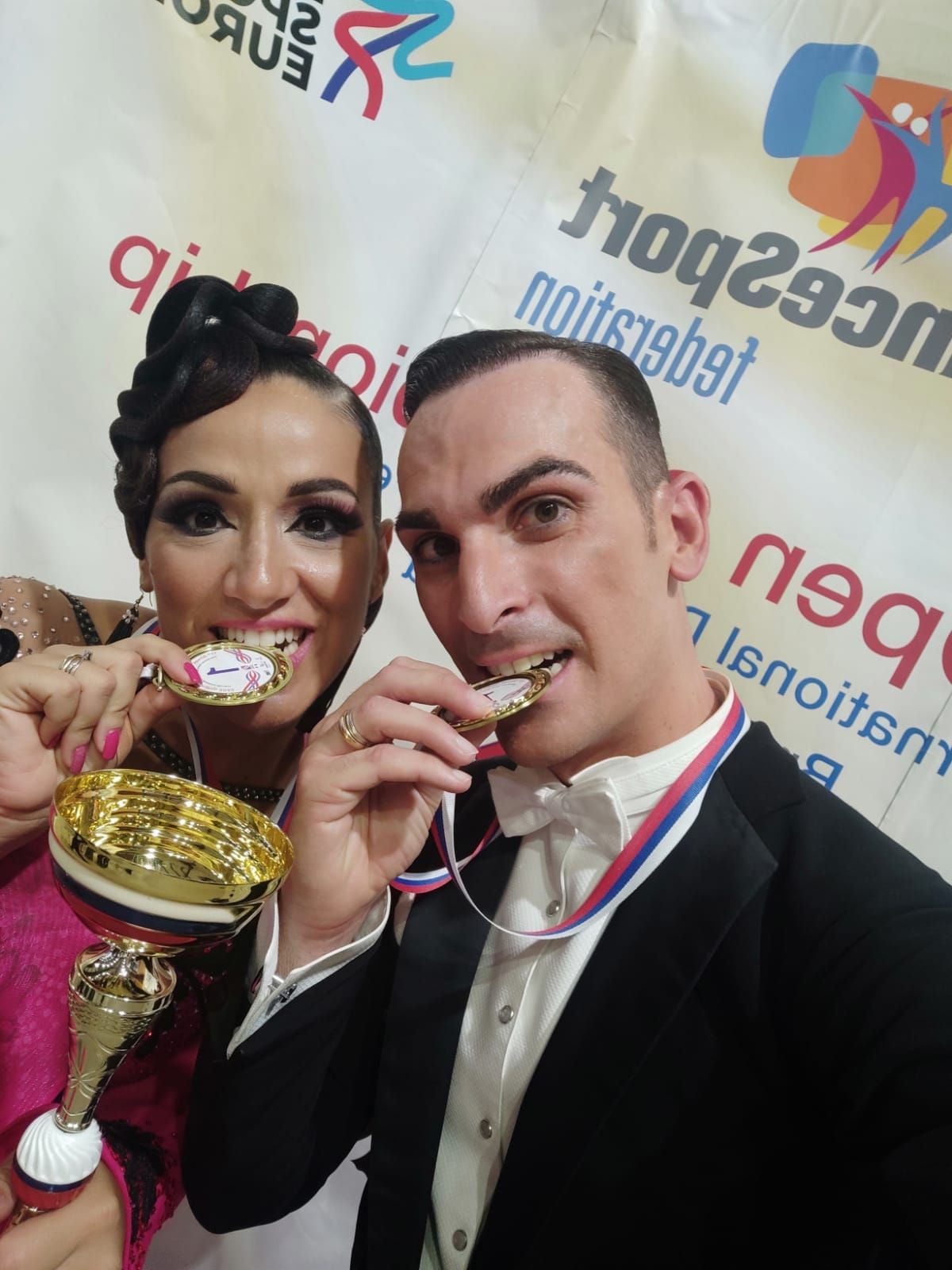 Una coppia che balla sorride con medaglie e trofei. Sono in abiti formali, in posa e in segno di festa.