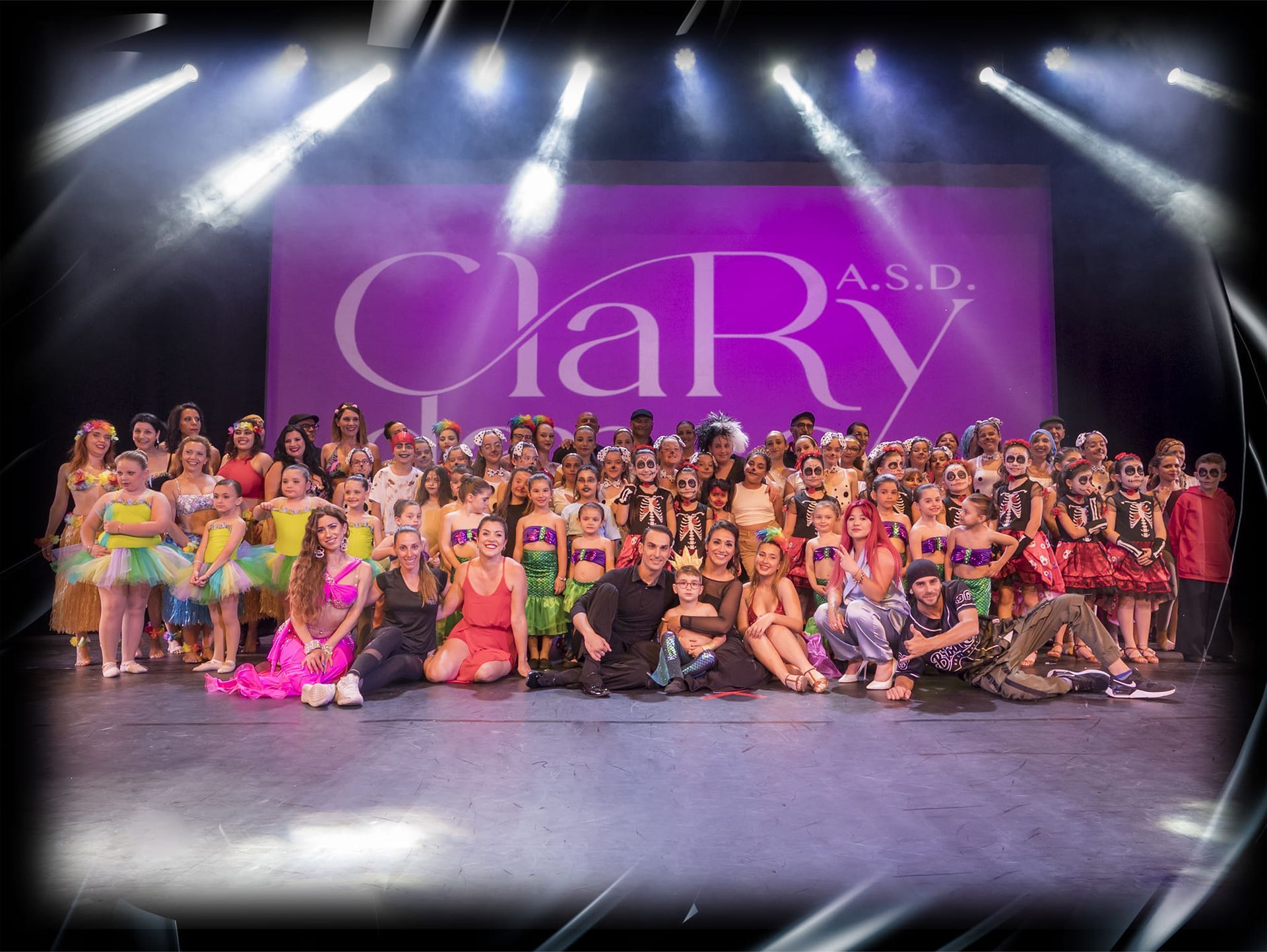 Un folto gruppo di ballerini in costumi colorati sul palco davanti allo sfondo 