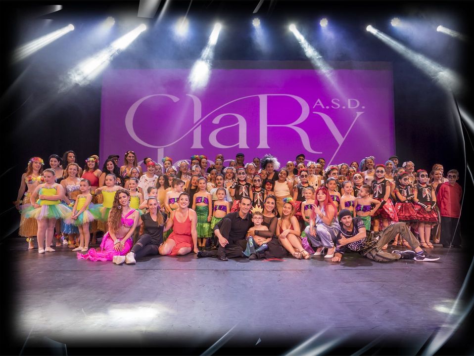 Un folto gruppo di ballerini posa sul palco davanti a uno sfondo viola con la scritta