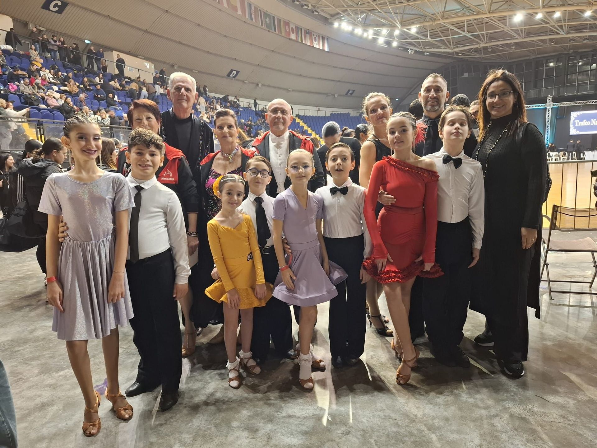 Gruppo di giovani ballerini in abiti formali, in posa con gli allenatori durante una gara, in un grande spazio al coperto.