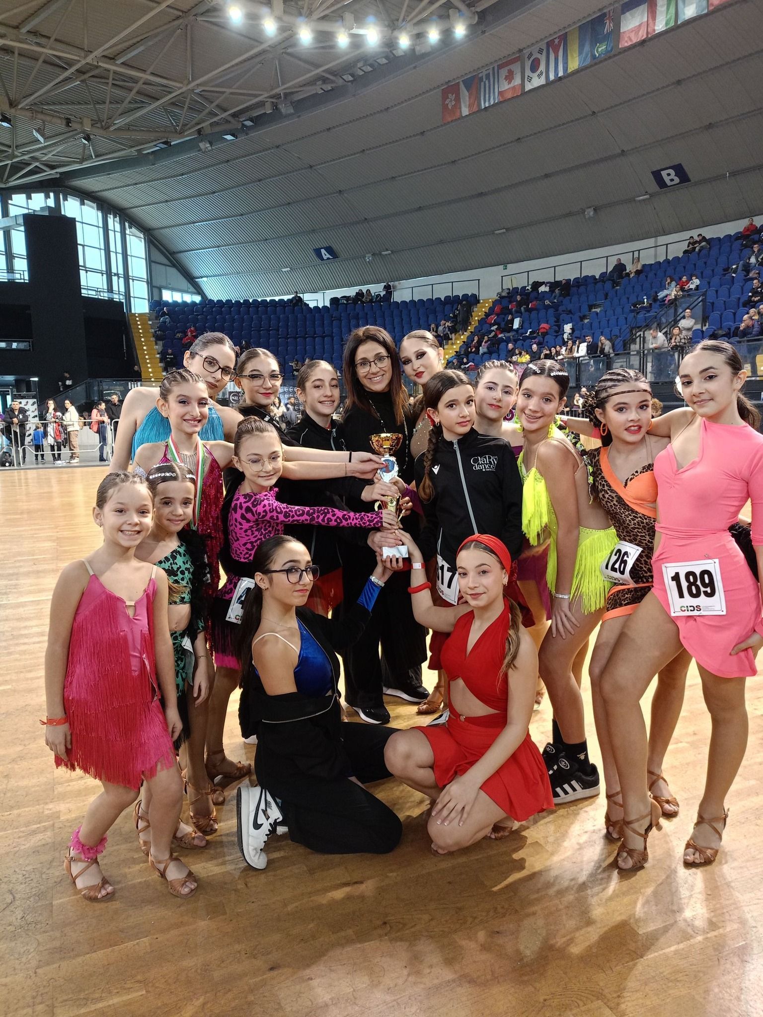 Una squadra di danza posa con un trofeo in un'arena, sorridendo e indossando costumi colorati.